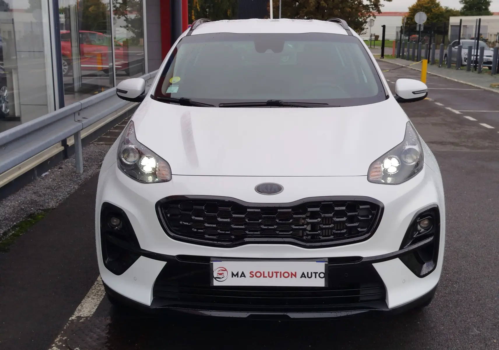 Vue frontale d'un KIA SPORTAGE blanc 2022 avec calandre noire et phares allumés sur parking.