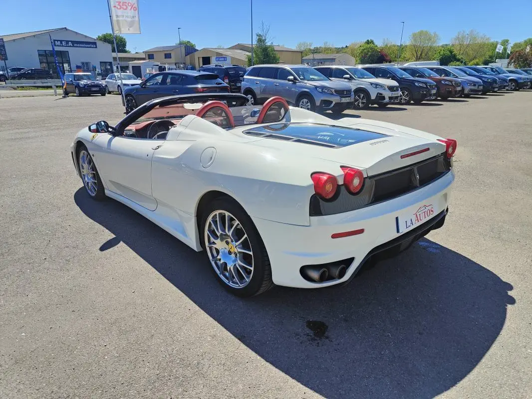 Ferrari F430 Spider blanc vue 3/4 arrière droit, capote ouverte et intérieur rouge visible, stationnée en extérieur.