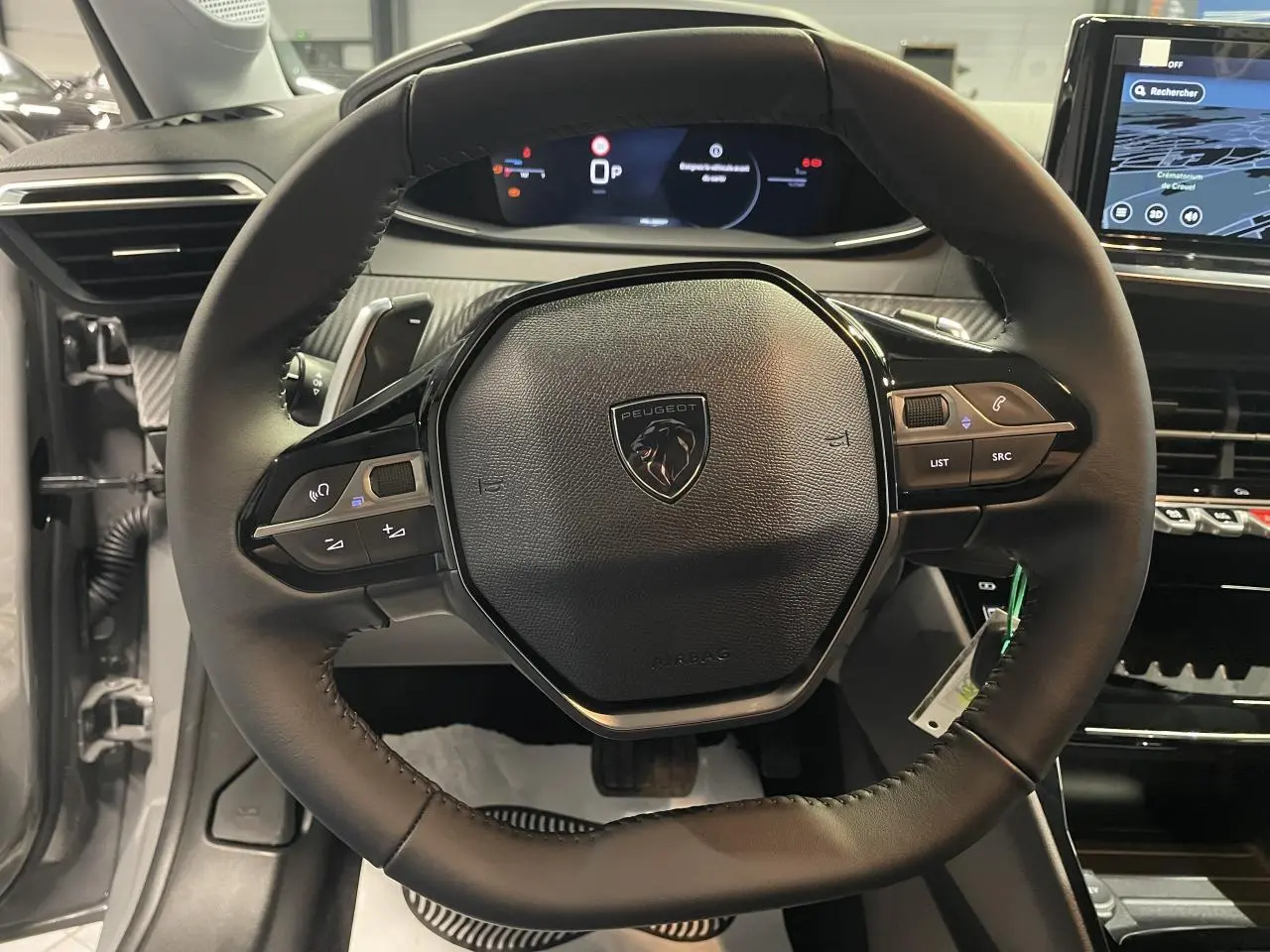 Vue rapprochée du volant cuir noir de la Peugeot 208 1.2i Hybrid 2025 avec commandes multimédia intégrées et tableau de bord numérique.