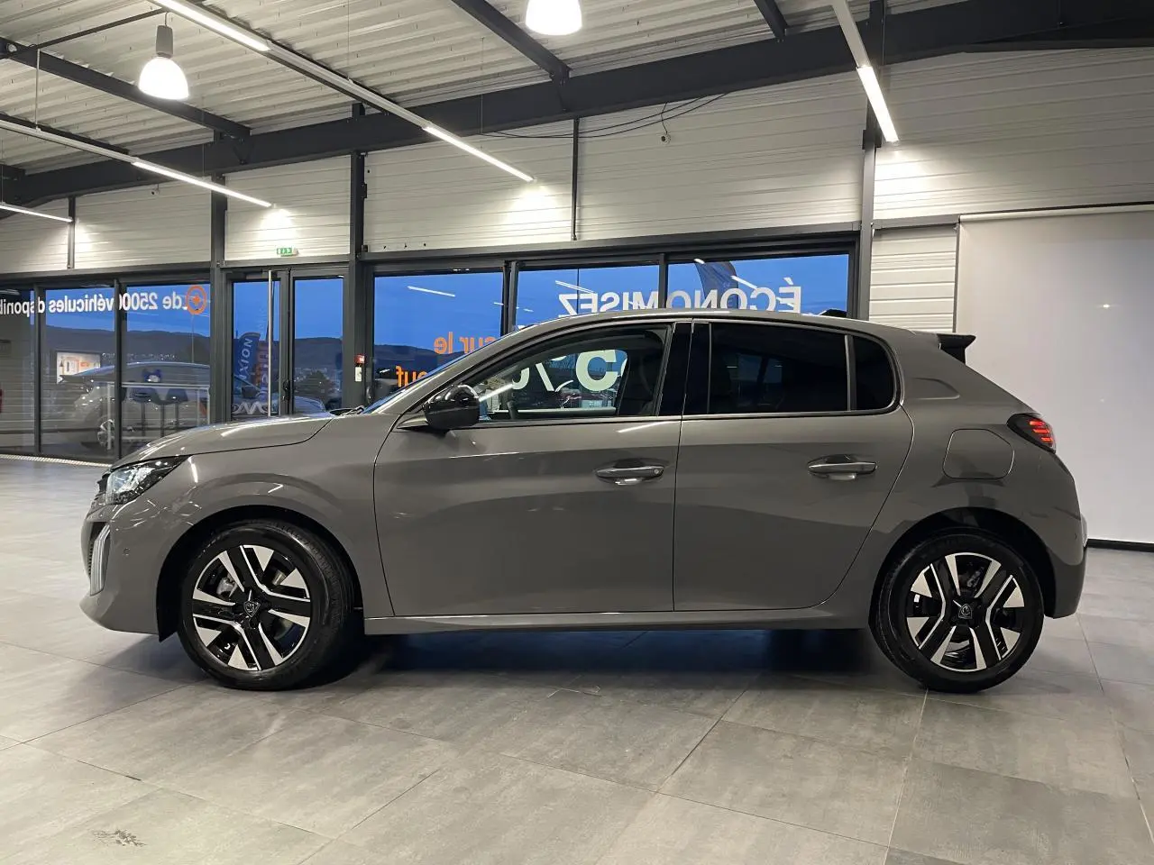 Vue de profil côté gauche d'une Peugeot 208 1.2i Hybrid gris selenium avec jantes alliage bi-tons en intérieur showroom.