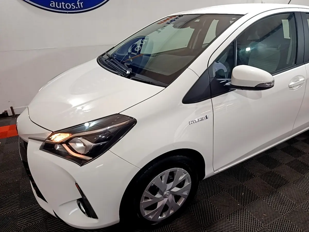 Vue 3/4 avant droit d'une Toyota Yaris blanche hybride avec phares allumés et logo Hybrid sur l'aile avant.
