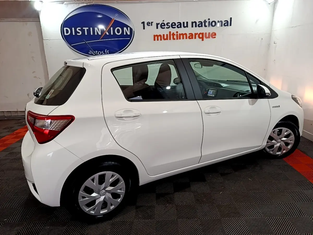 Profil droit de la Toyota Yaris hybride blanche 2019 en intérieur, avec jantes acier et logo Distinxion en arrière-plan.