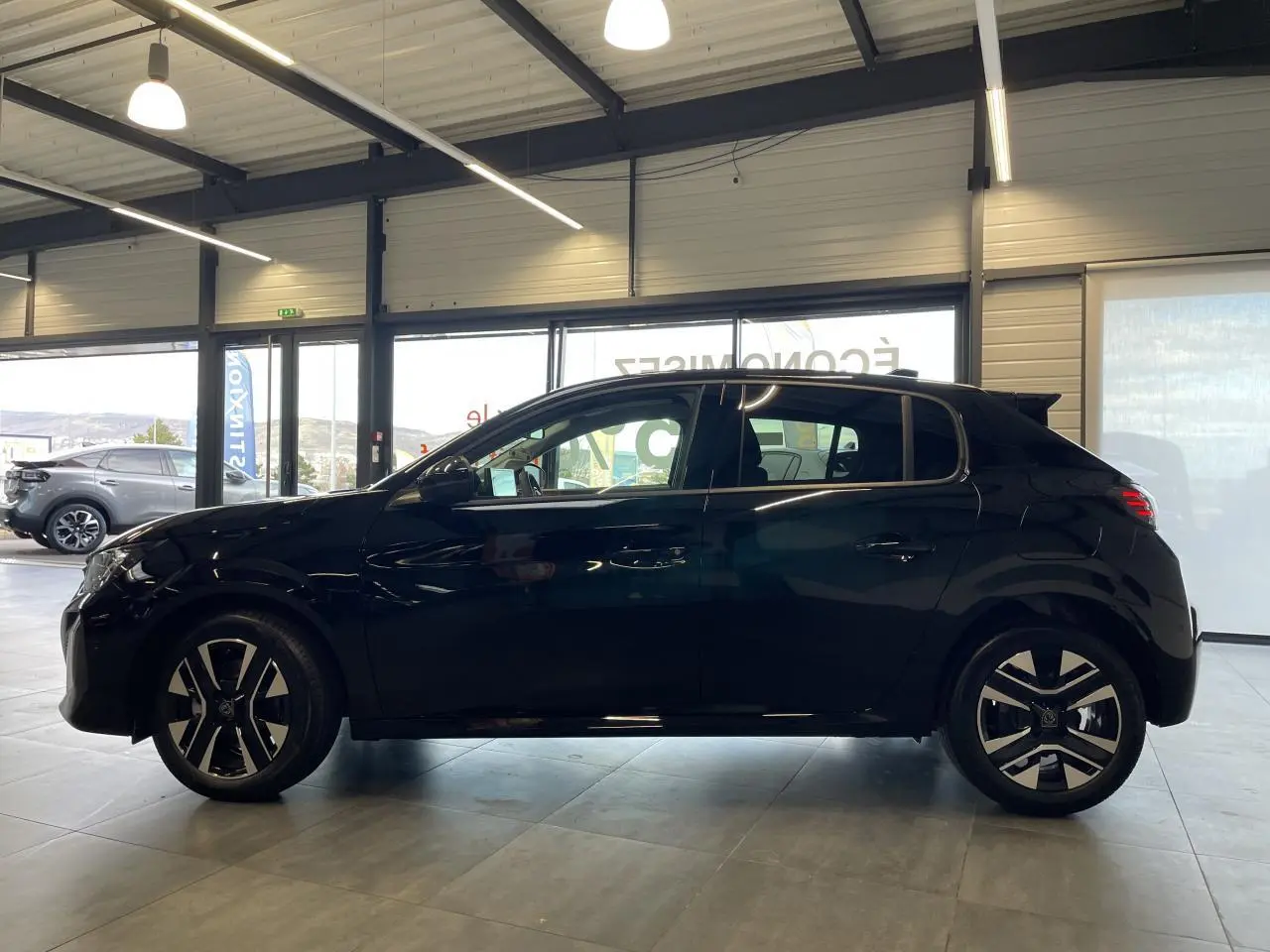 Profil côté gauche d'une Peugeot 208 noire 2025 avec jantes alliage bi-ton et vitres teintées en showroom.