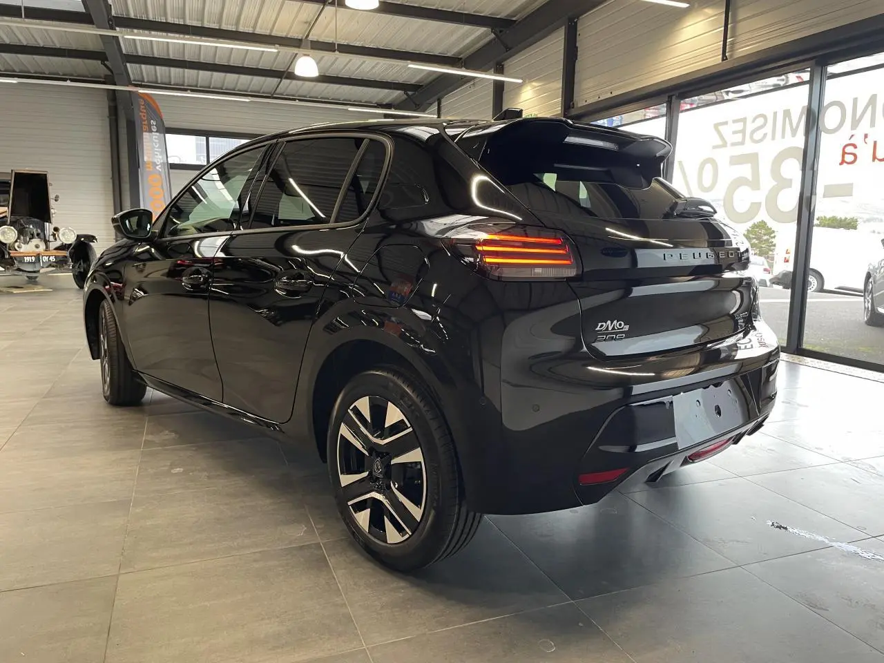 Peugeot 208 noire vue 3/4 arrière droit en intérieur, avec feux arrière allumés et jantes alliage bi-tons.