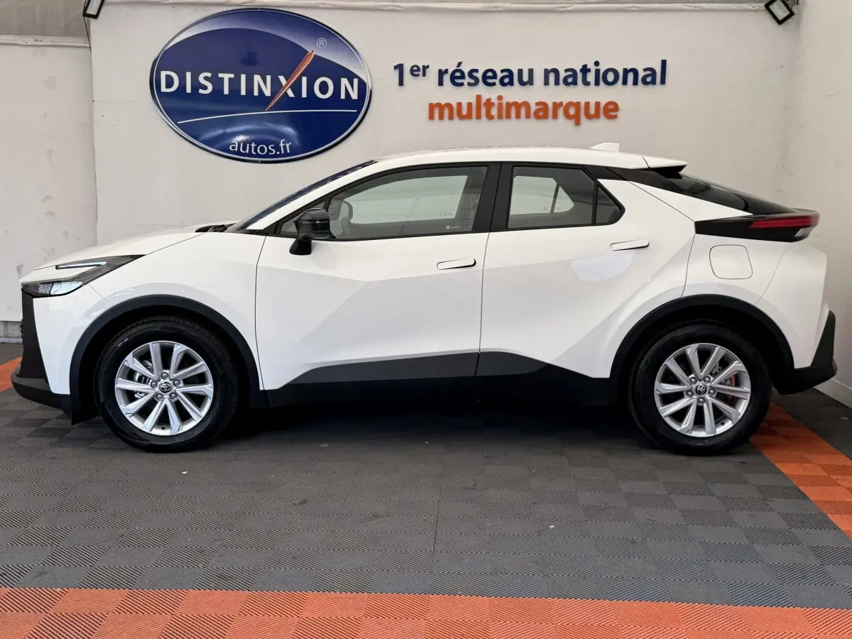 Profil côté gauche du Toyota C-HR blanc 2024 avec lignes anguleuses et jantes alliage visibles en intérieur showroom.