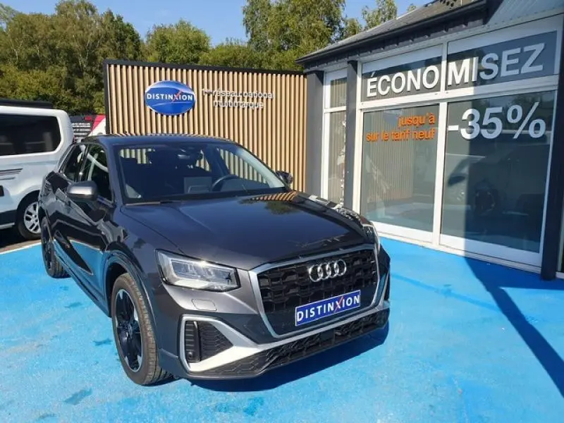 Audi Q2 30 TDI 116ch Design gris Manhattan métallisé vue 3/4 avant droit sur parking bleu devant vitrine promotionnelle.