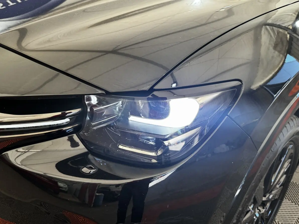 Gros plan sur le phare avant droit allumé d’un Mazda CX-60 noir, mettant en valeur la calandre et la carrosserie brillante.