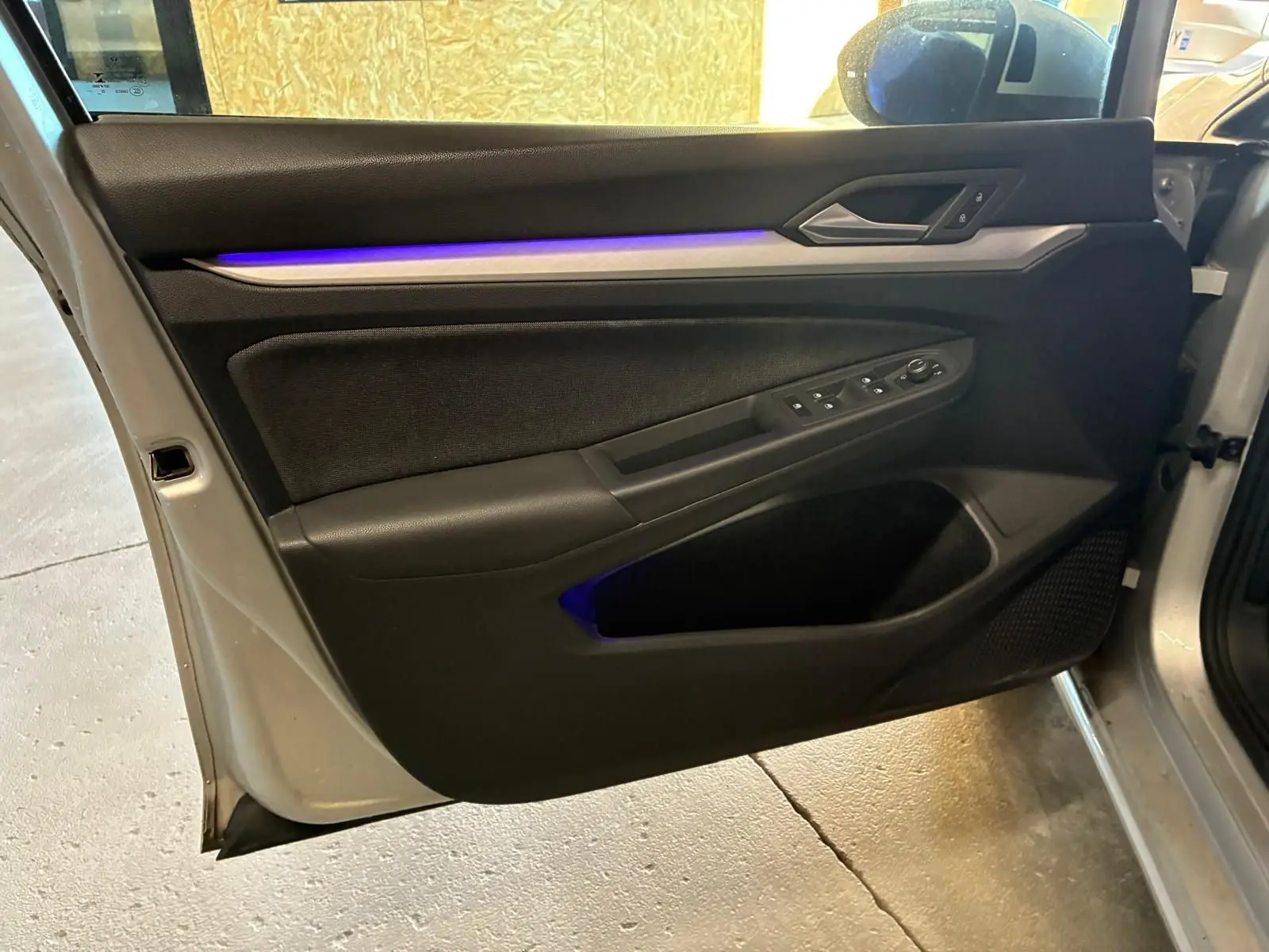 Vue intérieure de la porte avant gauche d'une Volkswagen Golf gris clair 2023, avec éclairage d'ambiance bleu et commandes électriques.