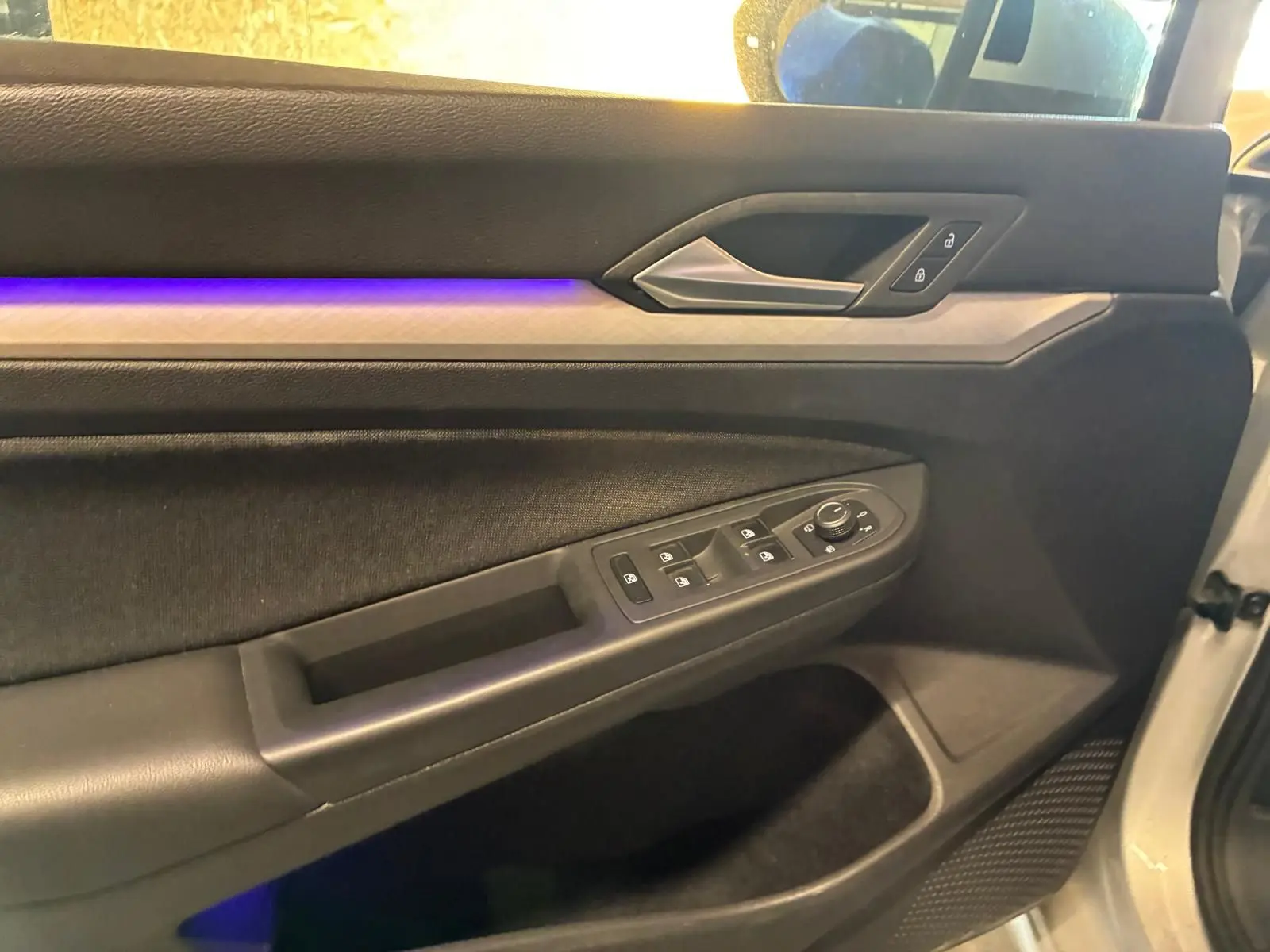 Vue intérieure côté conducteur de la porte gris clair d'une Volkswagen Golf 2023 avec éclairage d'ambiance violet et commandes électriques.