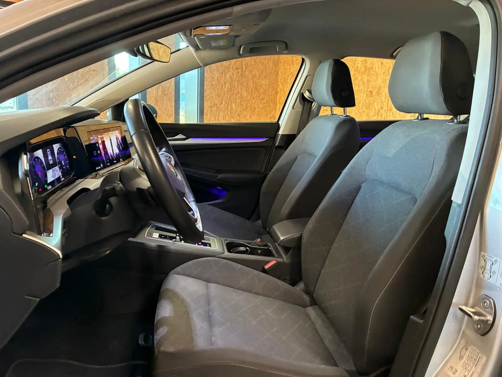 Vue intérieure côté conducteur de la Volkswagen Golf gris clair 2023, avec tableau de bord numérique et éclairage d’ambiance violet.