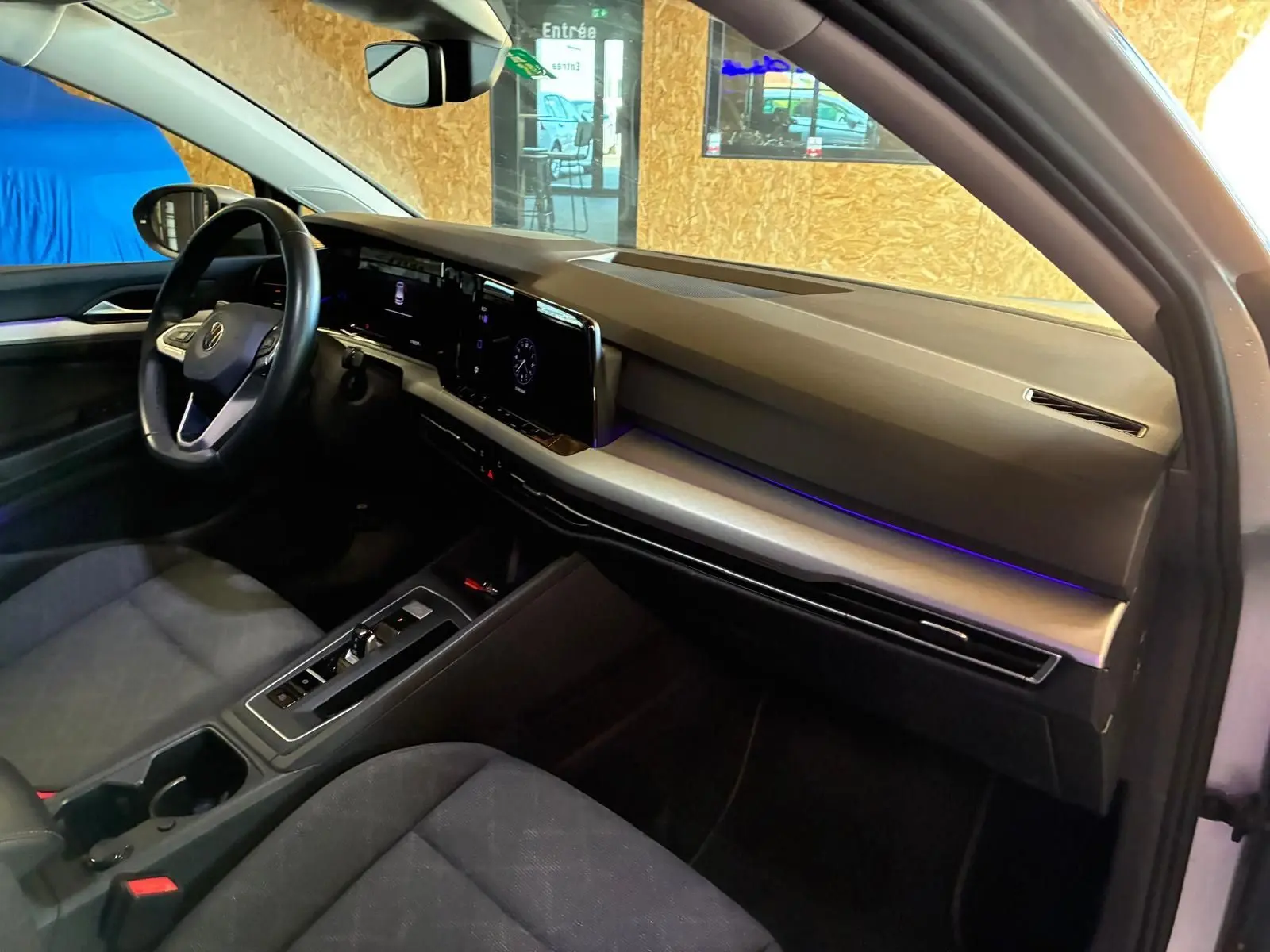 Vue intérieure côté conducteur de la Volkswagen Golf 2023 gris clair, avec tableau de bord digital et éclairage d'ambiance violet.