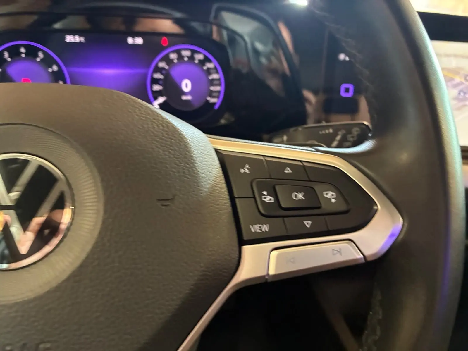 Gros plan sur le volant côté droit d’une Volkswagen Golf gris clair 2023, avec commandes intégrées et tableau de bord numérique violet.
