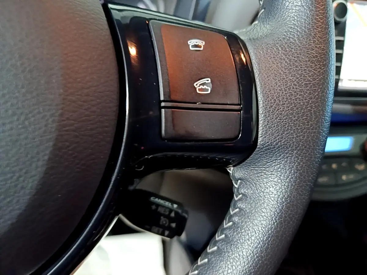 Gros plan sur les commandes noires du volant cuir et alu de la Toyota Yaris blanche, version hybride 2020.