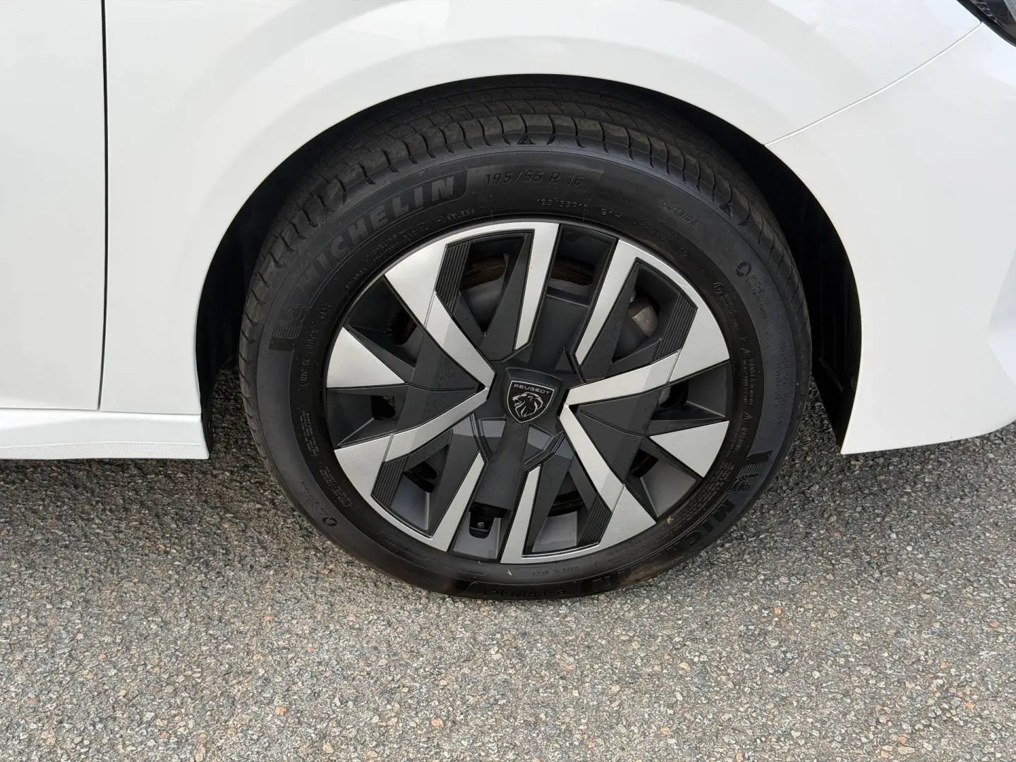 Gros plan sur la roue avant gauche blanche Okénite d’une Peugeot 208 1.2 Hybrid avec jante au design géométrique noir et argent.