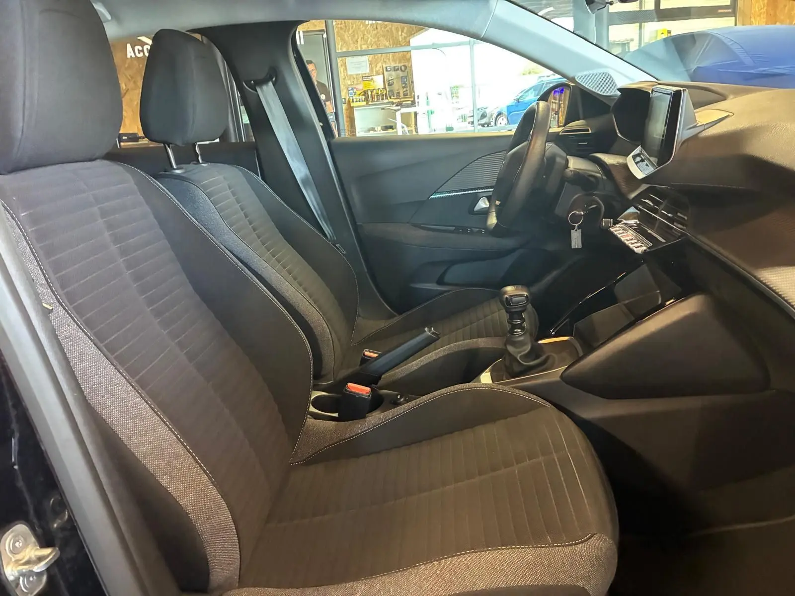 Vue intérieure côté conducteur de la Peugeot 208 noire, mettant en valeur les sièges en tissu et la console avec boîte manuelle.