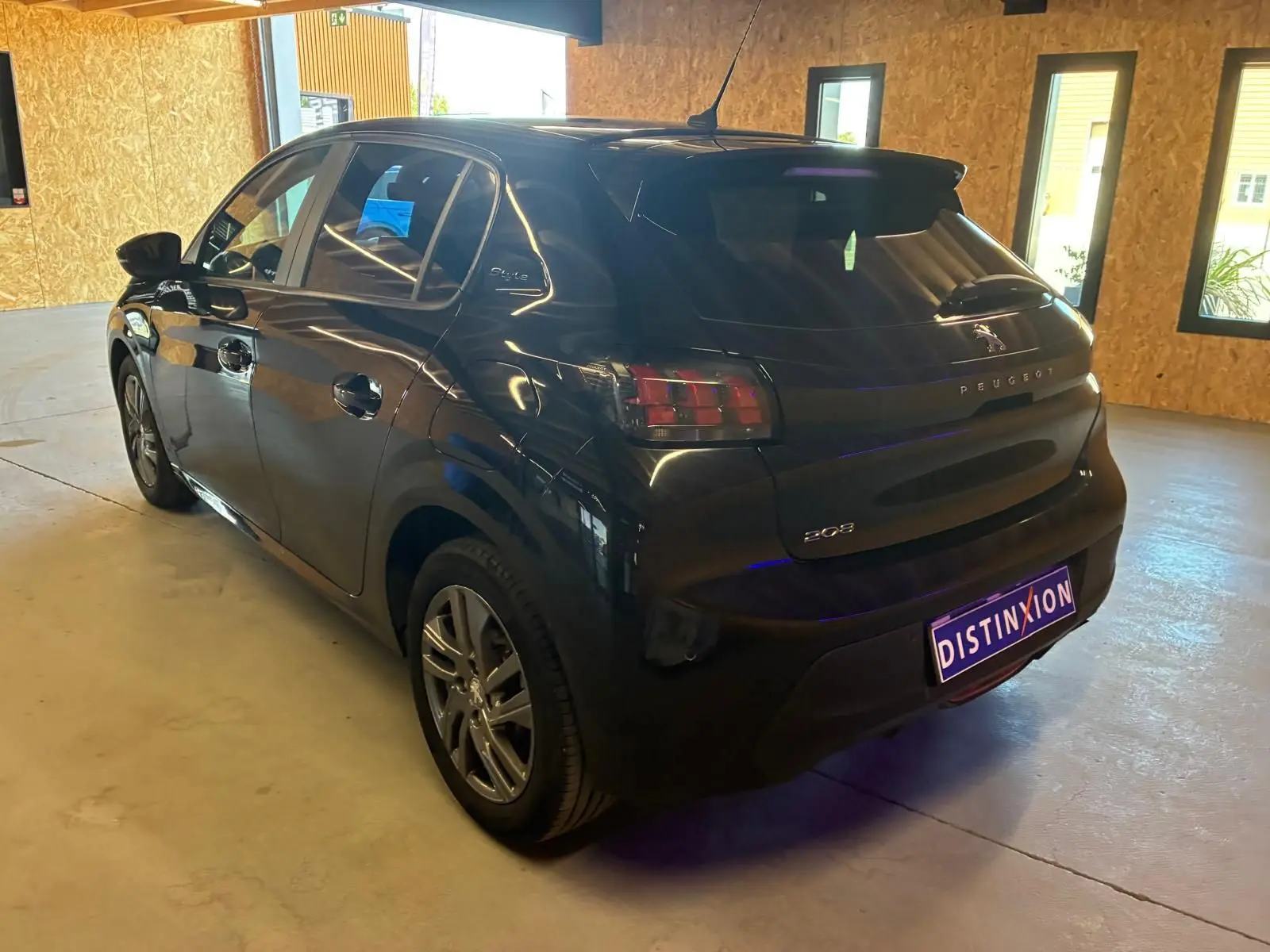 Vue 3/4 arrière droite d'une Peugeot 208 noire 5 portes, stationnée en intérieur avec plaques Distinxion visibles.