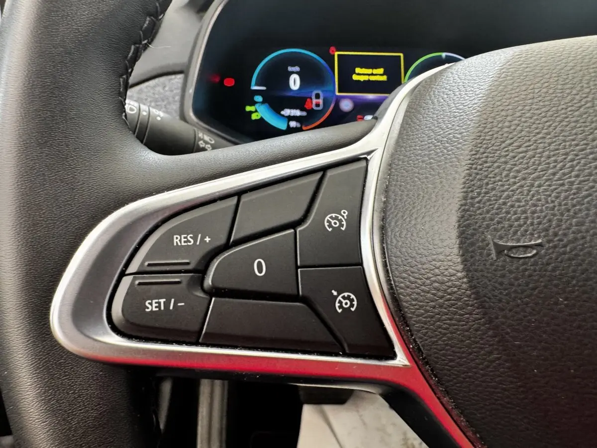 Gros plan sur les commandes du régulateur de vitesse à gauche du volant cuir noir de la Renault ZOE blanche, tableau de bord numérique en arrière-plan.