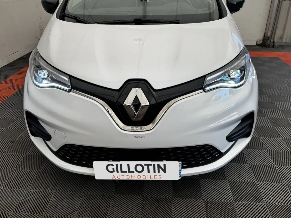 Vue avant rapprochée d'une Renault ZOE blanche avec phares LED allumés et logo Renault bien visible.