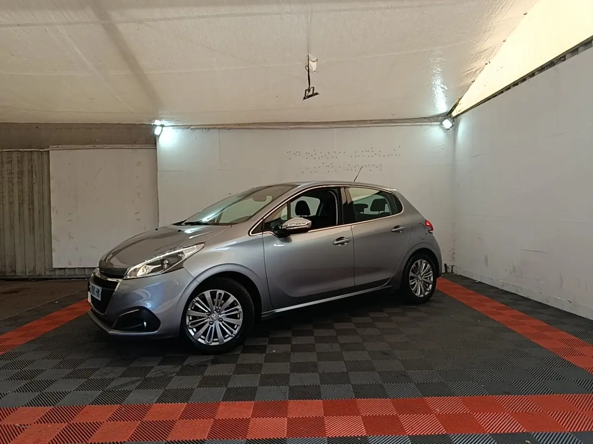 Peugeot 208 Puretech 82 Allure gris clair vue 3/4 avant droit dans un garage éclairé.