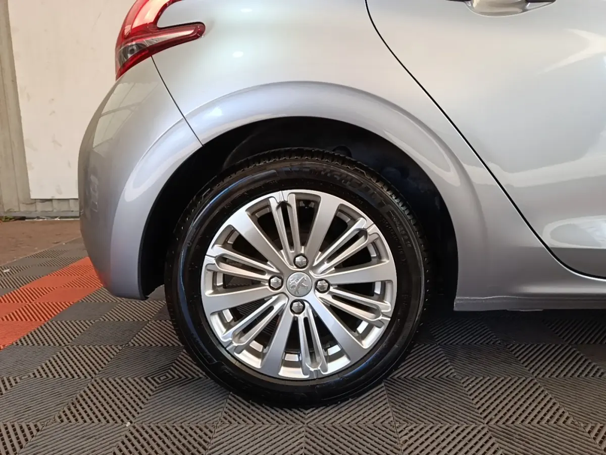 Gros plan sur la roue arrière droite d'une Peugeot 208 gris clair, avec jante alliage et feu arrière partiel visible.