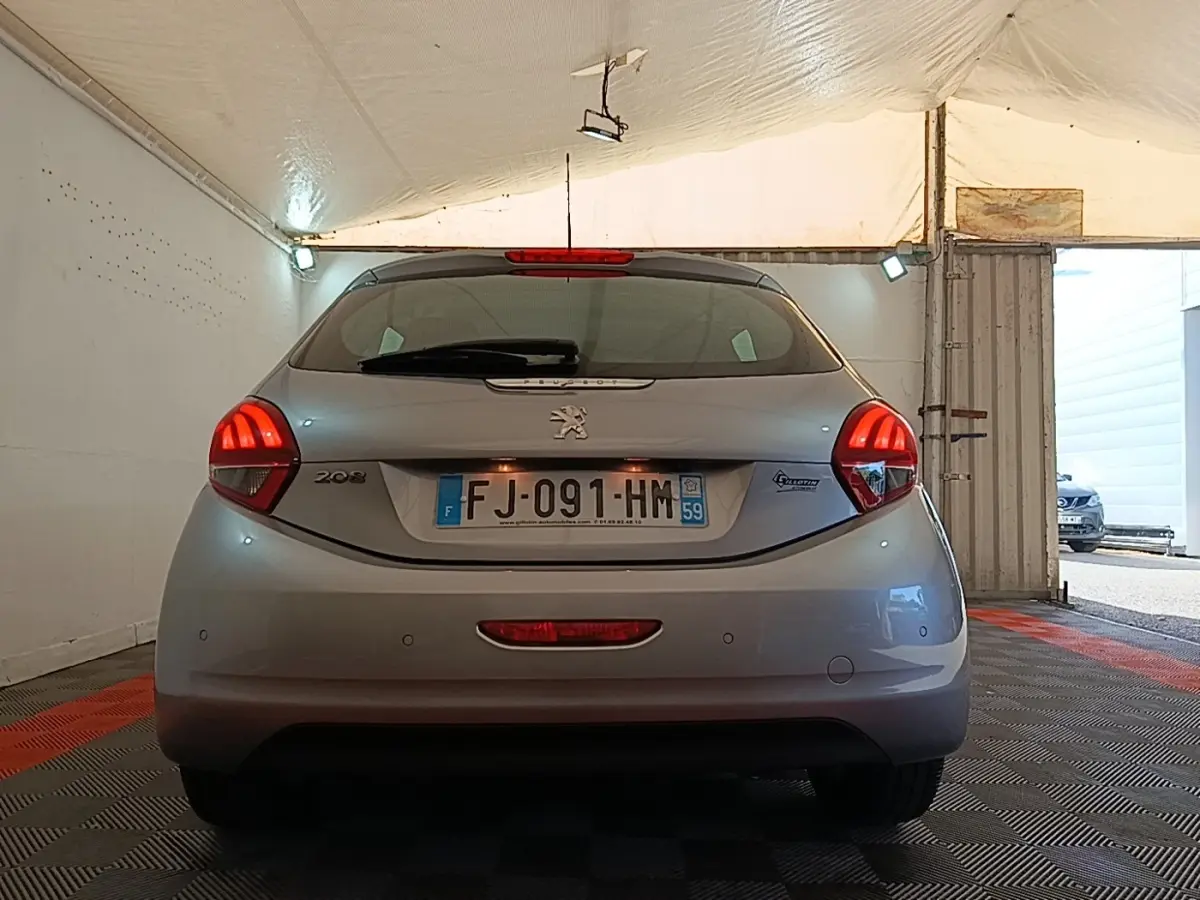 Vue arrière d'une Peugeot 208 gris clair garée en intérieur, avec feux arrière allumés et plaque d'immatriculation visible.