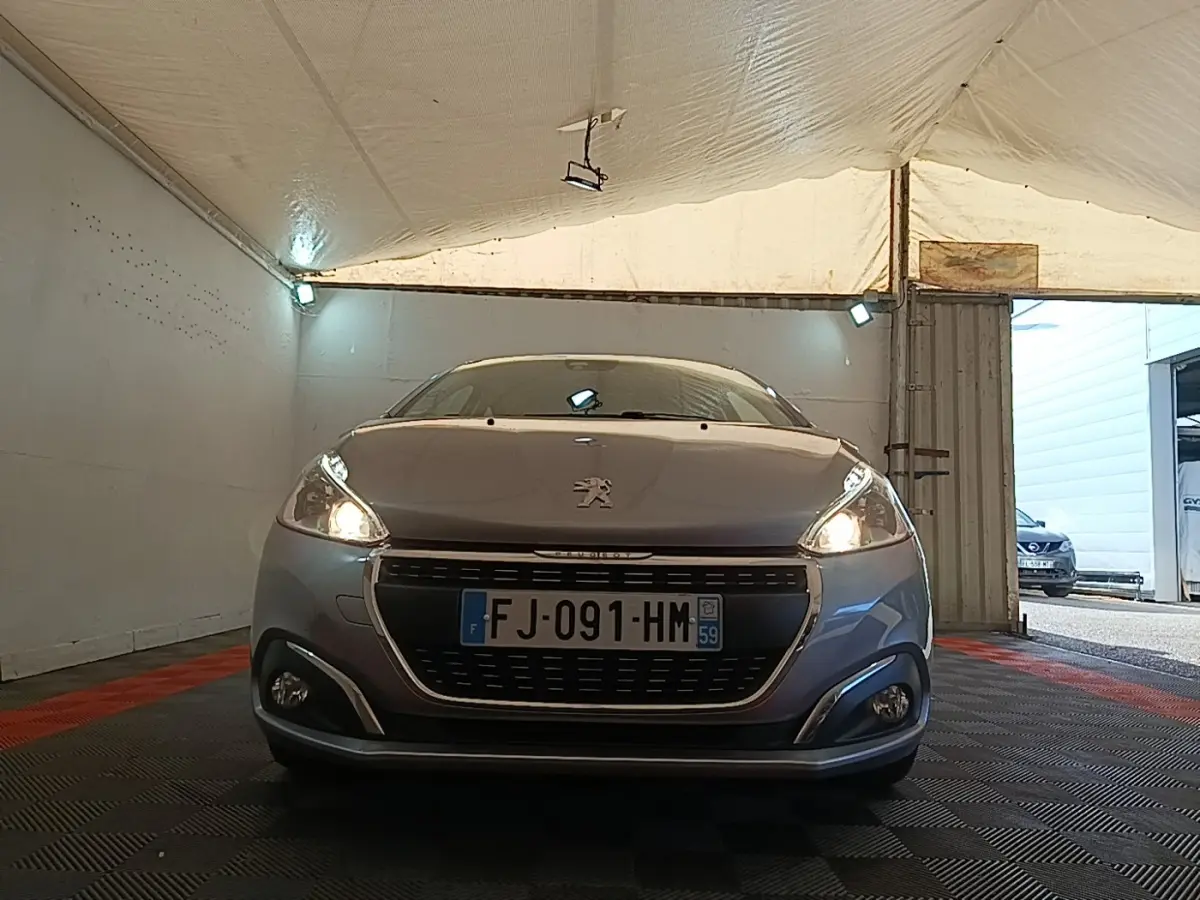 Vue frontale d'une Peugeot 208 gris clair avec phares allumés dans un garage couvert.