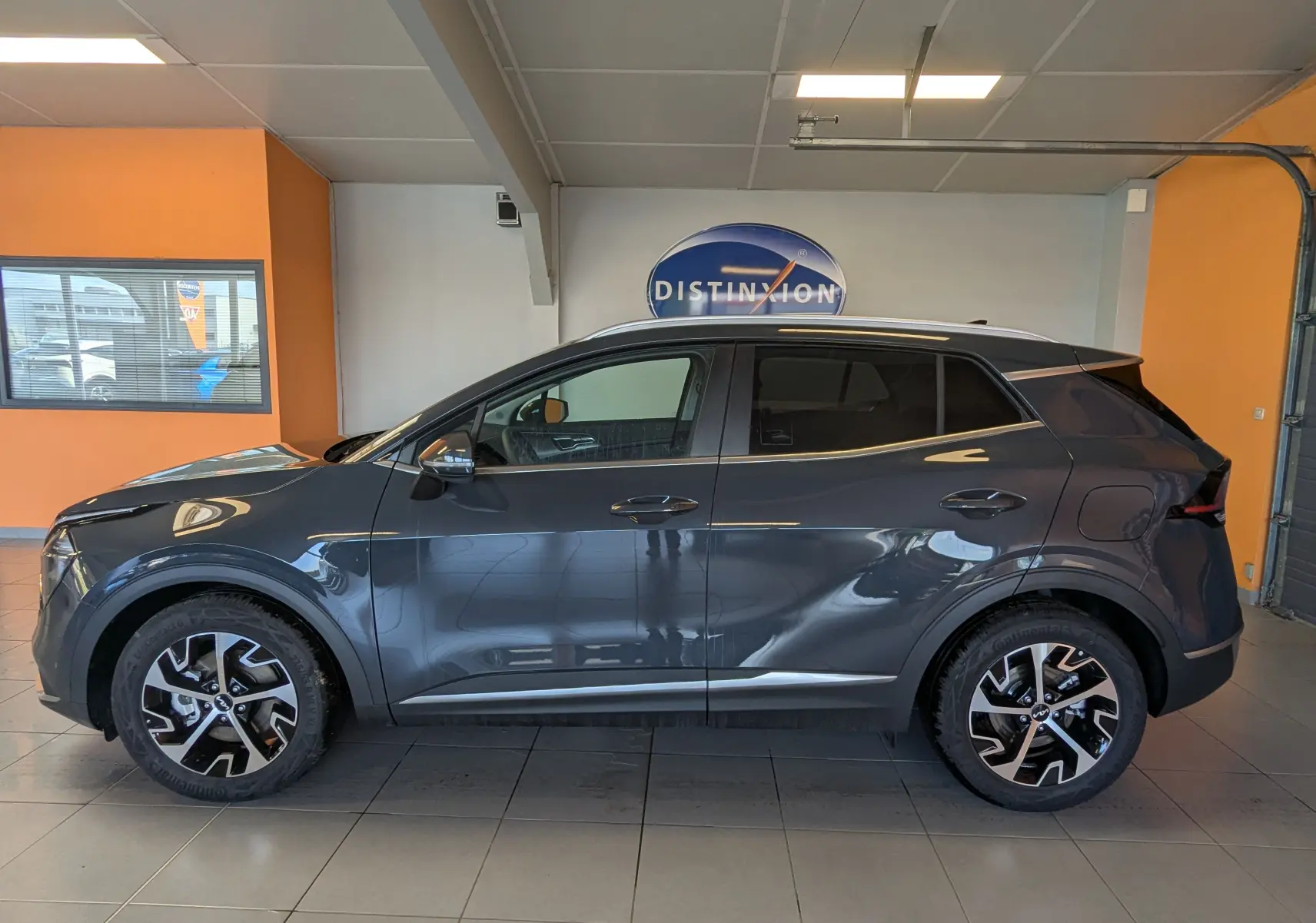 Profil côté gauche du KIA SPORTAGE gris eclipse MY25 avec jantes alliage 18'', barres de toit et vitres teintées en showroom.