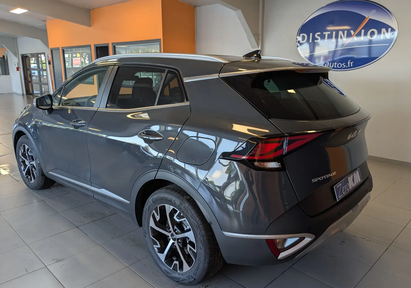 Vue 3/4 arrière droite du KIA SPORTAGE gris eclipse MY25 avec jantes alliage 18'' et barres de toit en intérieur showroom.