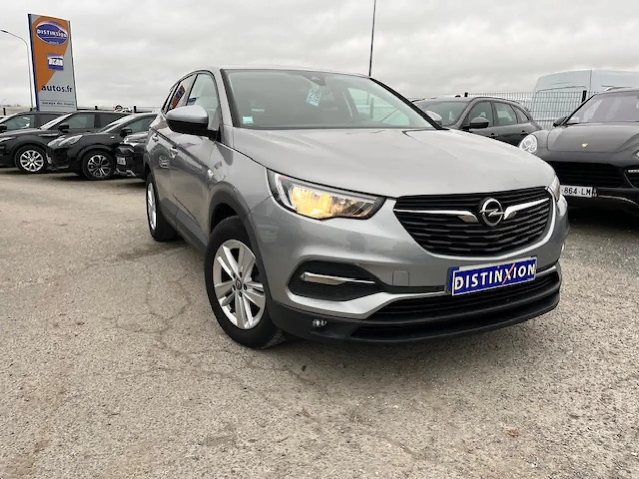 Opel Grandland X gris clair vu en 3/4 avant droit avec phare allumé et jantes alliage 17 pouces sur parking.