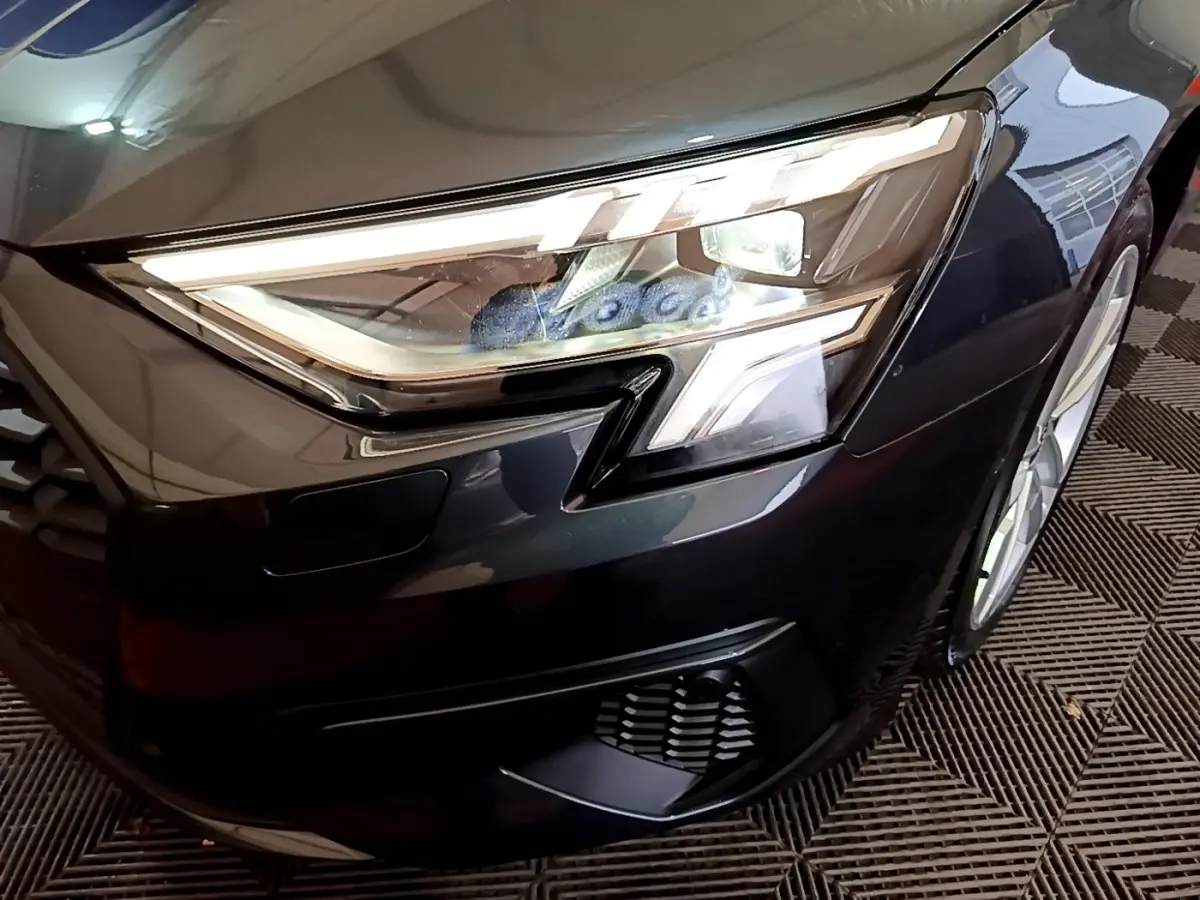 Gros plan sur le phare avant droit d'une Audi A3 gris foncé avec détails LED et partie de la calandre visible.
