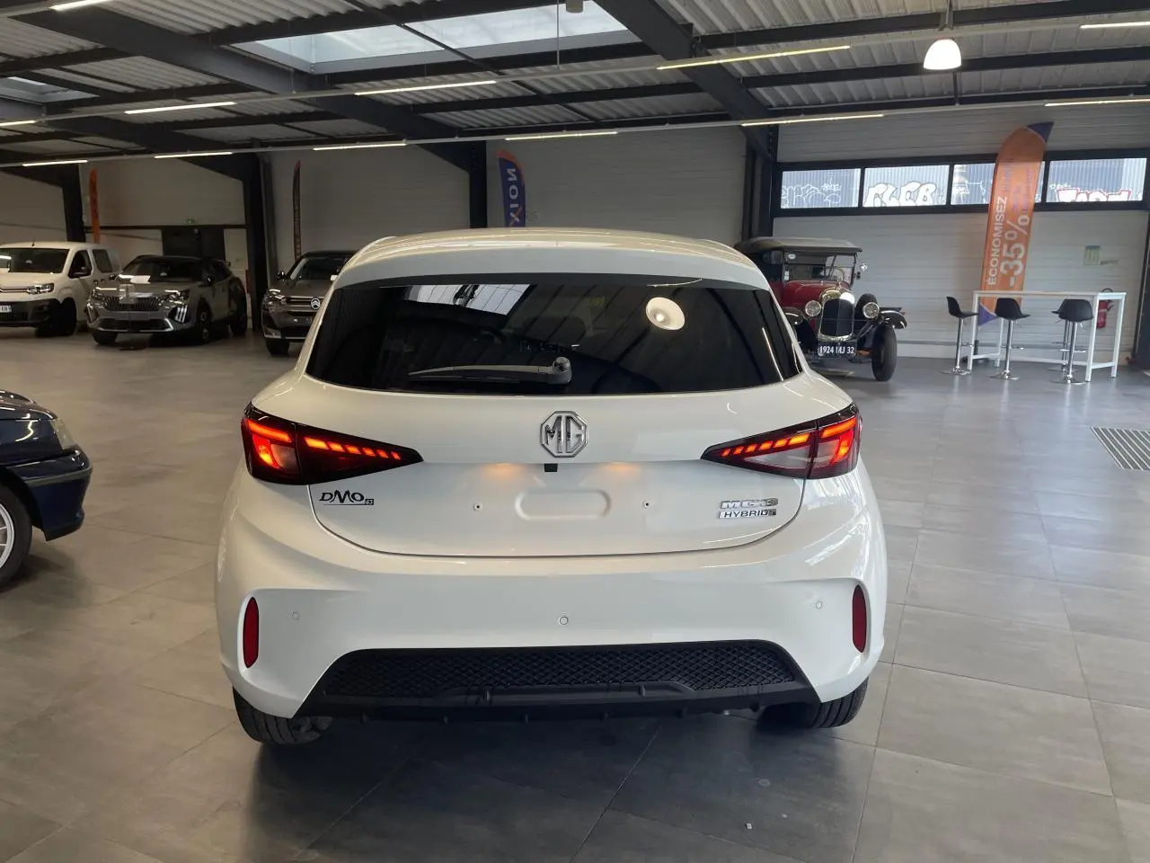 Vue arrière d'une MG3 1.5 Hybrid+ blanche avec feux LED allumés dans un showroom intérieur.