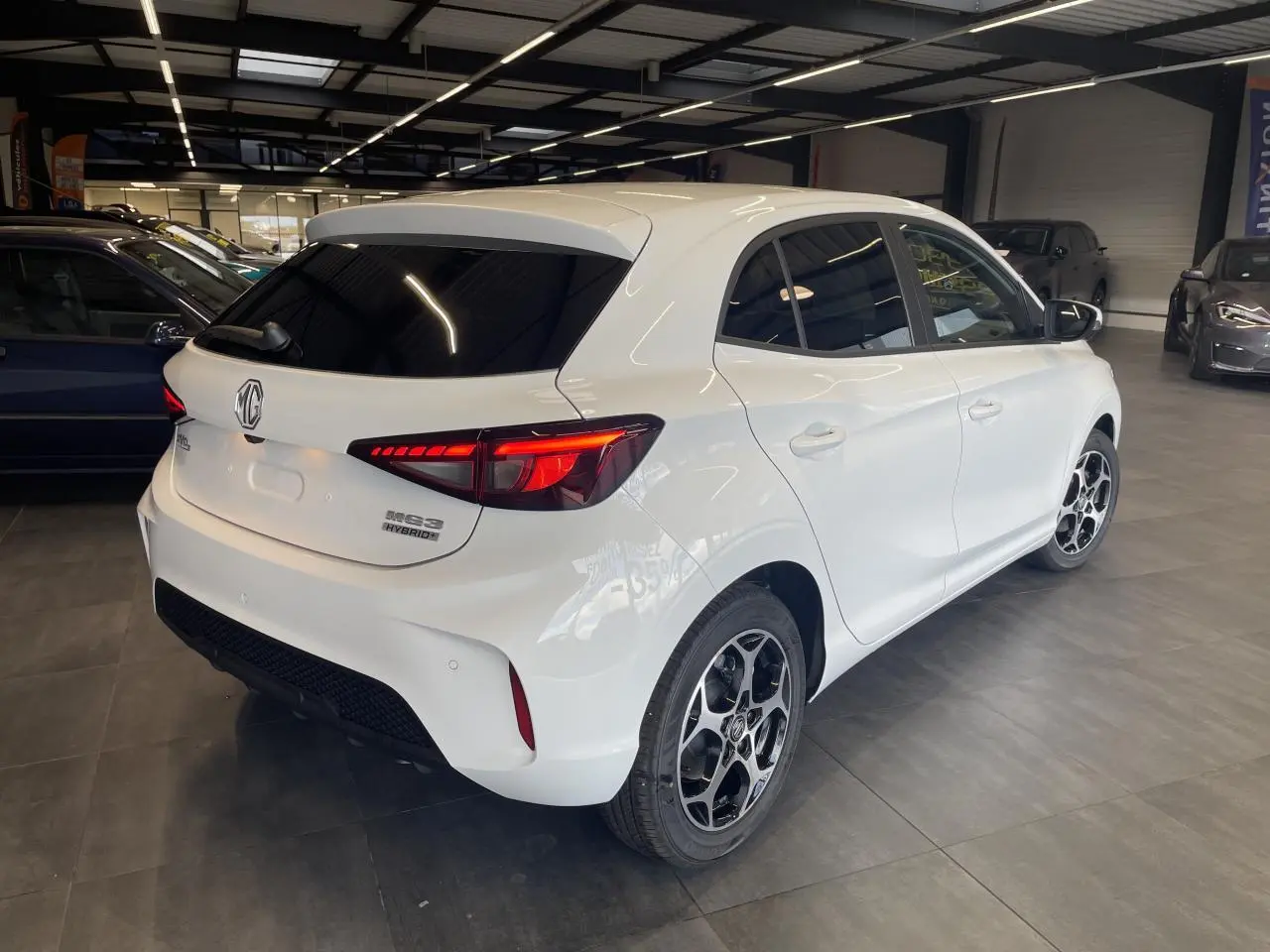 Vue 3/4 arrière droite d'une MG3 blanche avec feux LED allumés et jantes alliage noires en showroom.