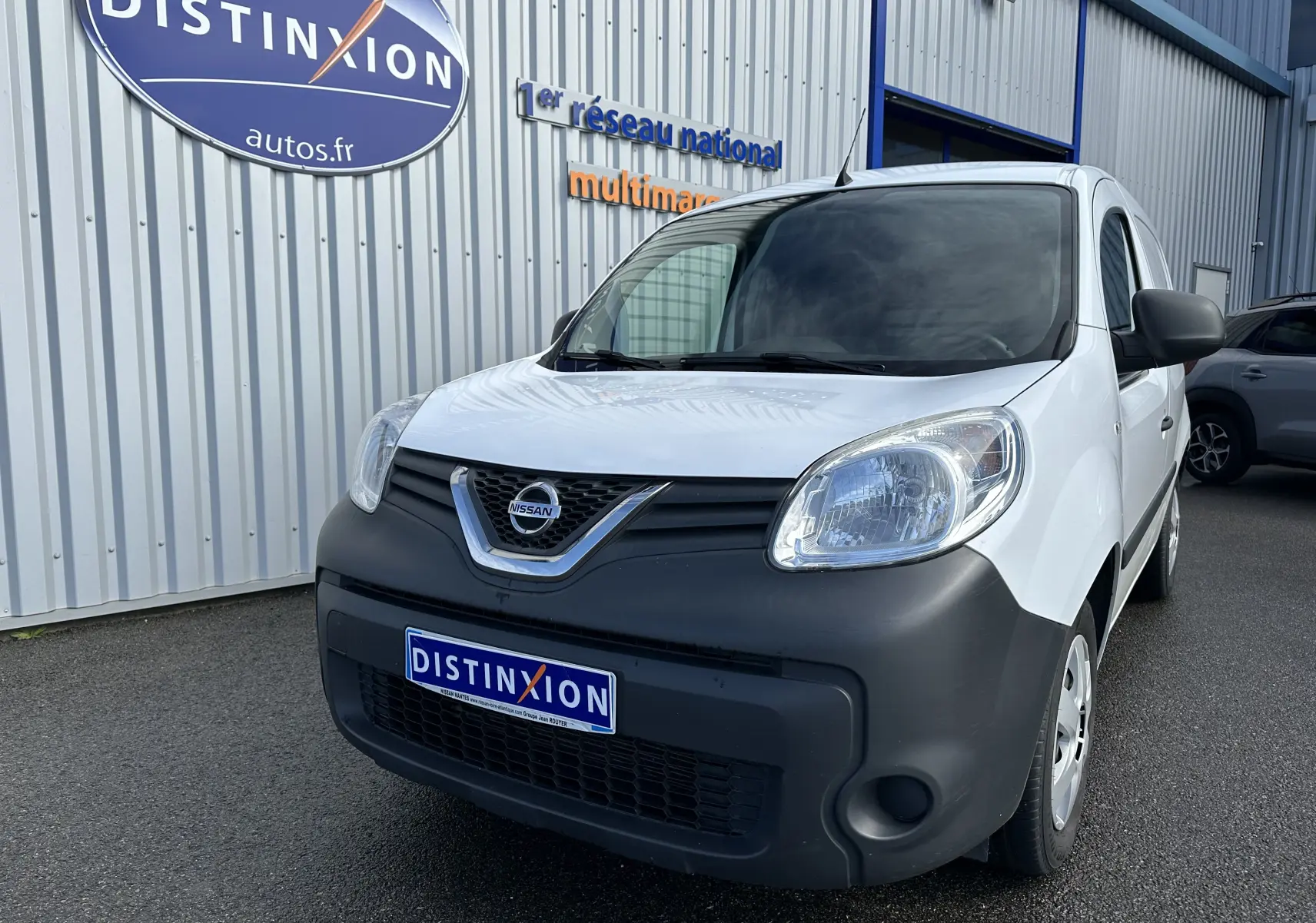 Nissan NV250 blanc en 3/4 avant droit, avec pare-chocs noir mat et logo Nissan visible sur la calandre.