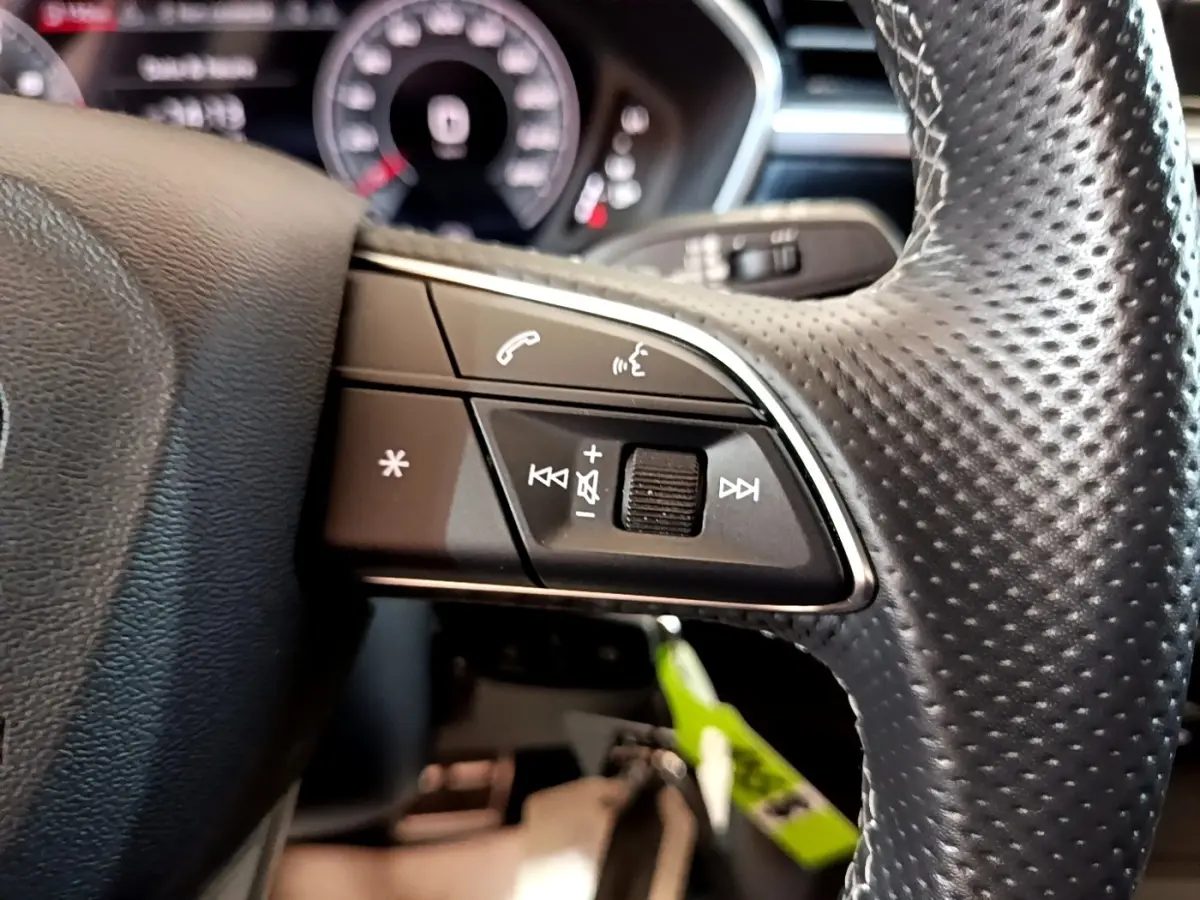 Gros plan sur les commandes gauche du volant cuir perforé de l'Audi Q3 gris foncé, avec tableau de bord numérique flou en arrière-plan.