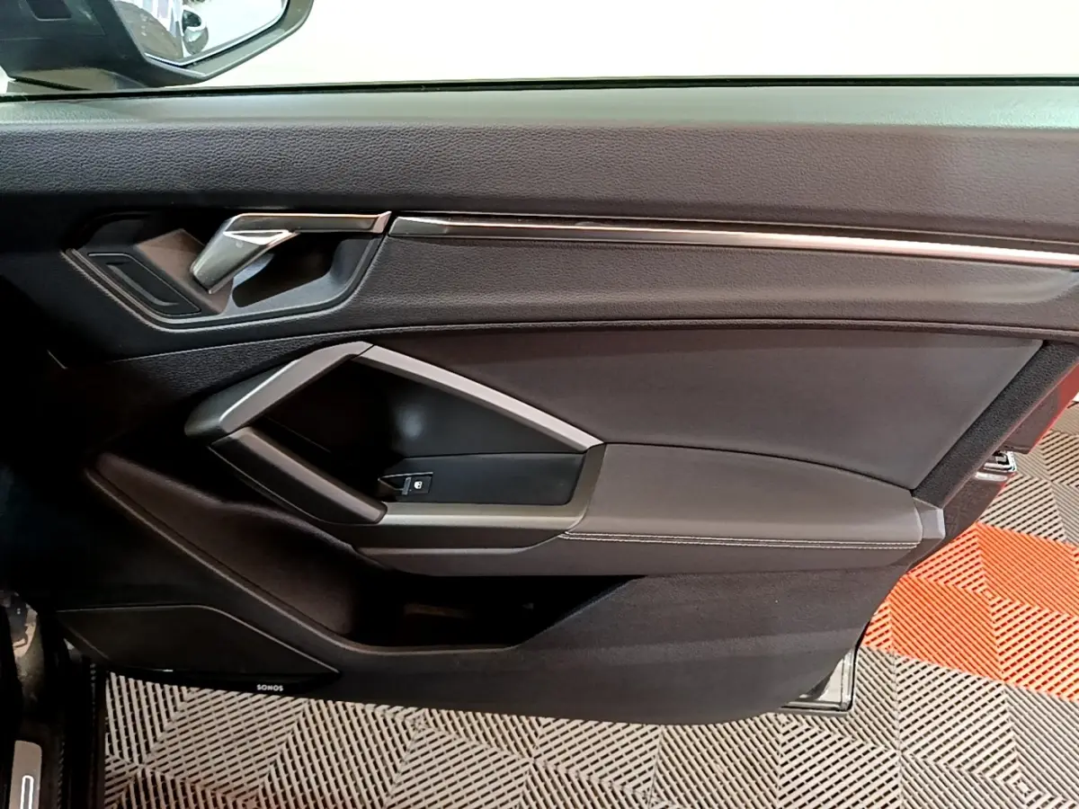 Vue intérieure côté gauche de la porte avant d’un Audi Q3 gris foncé avec poignée argentée et commande de vitre.