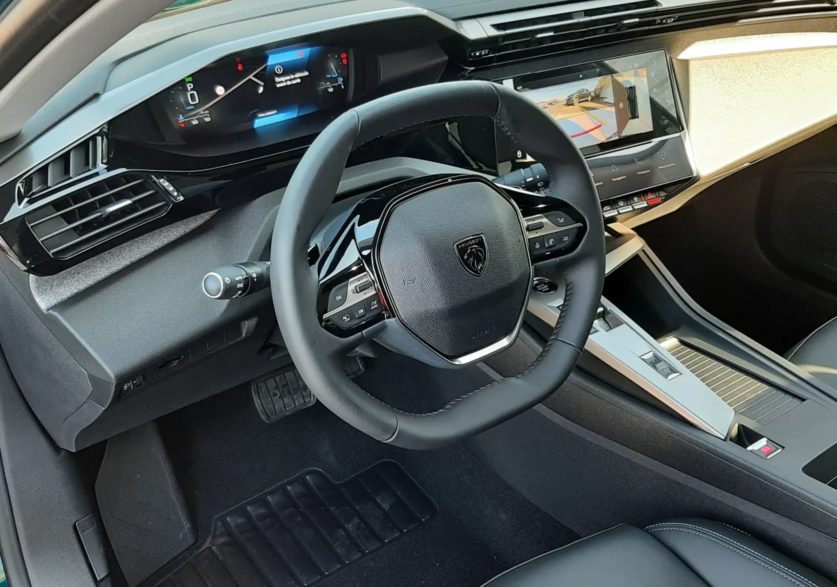 Vue intérieure côté conducteur de la Peugeot 308 2026, volant compact, tableau de bord numérique et écran central affichant la caméra 360°.