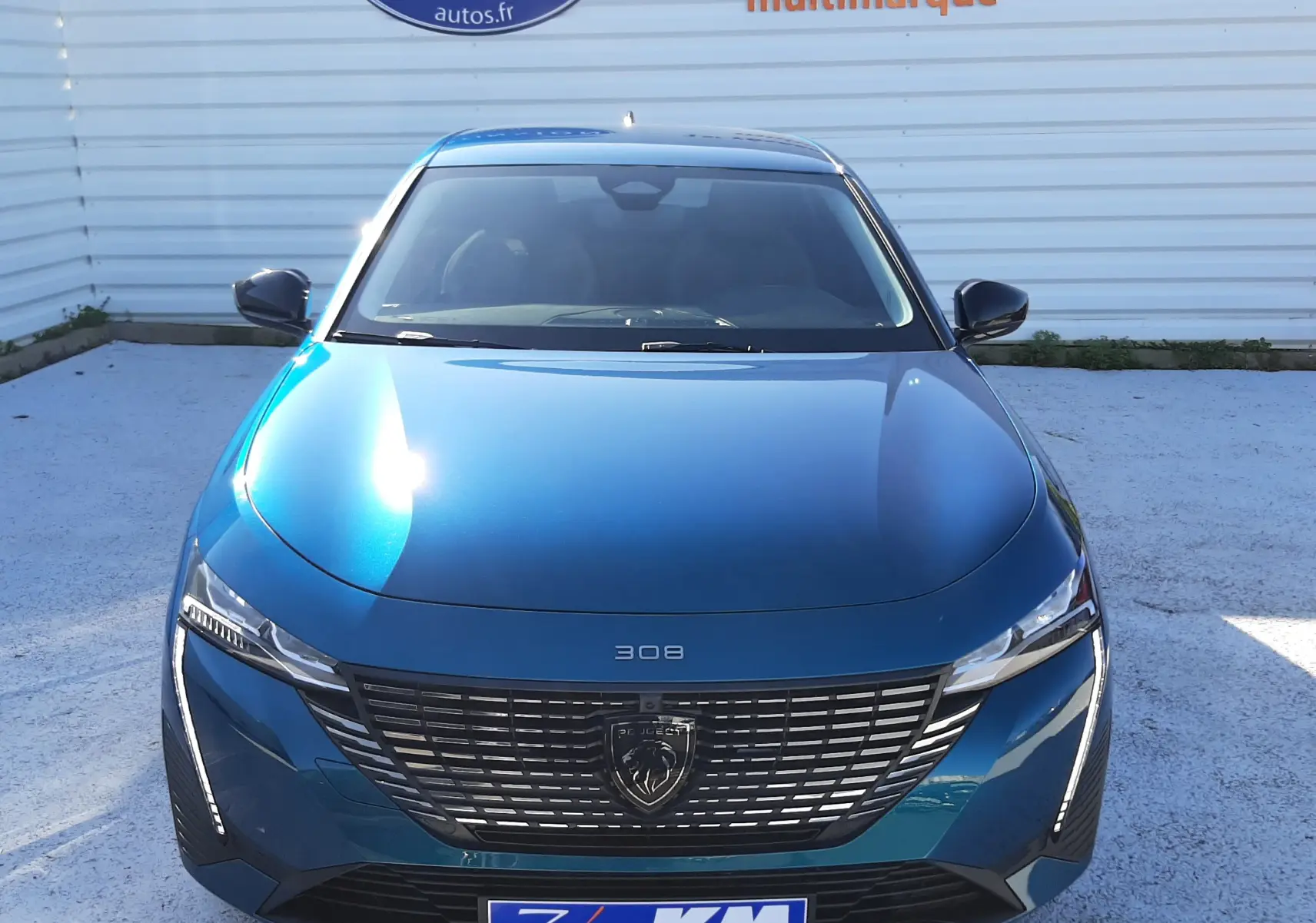 Vue frontale d'une Peugeot 308 Bleu Obsession 2026 avec calandre noire et logo lion distinctif, stationnée devant un mur blanc.