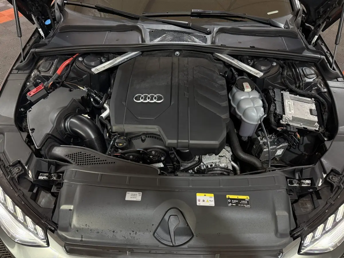 Vue plongeante sur le moteur noir d'une Audi A4 gris foncé, avec capot ouvert et logo Audi visible.