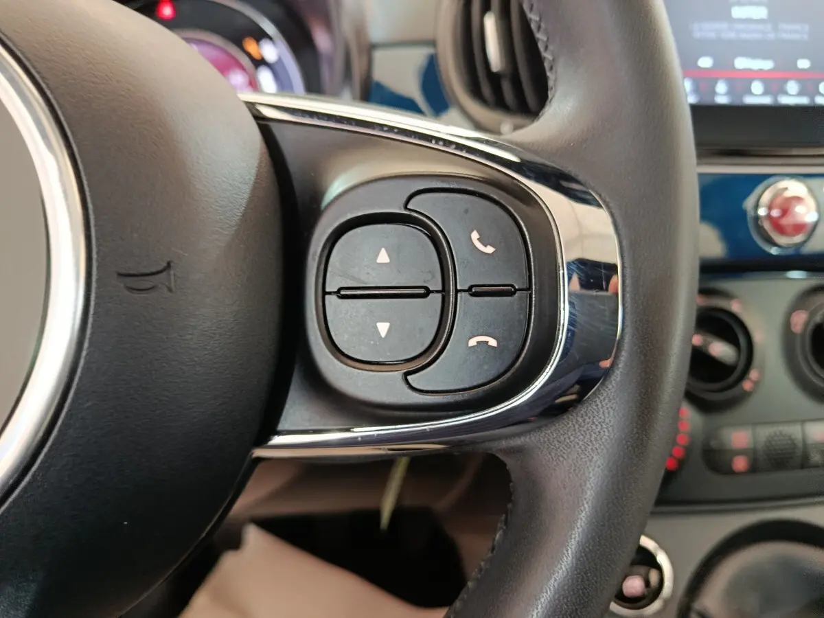 Gros plan sur les commandes du volant cuir noir de la Fiat 500 Hybrid bleu, avec tableau de bord et écran tactile en arrière-plan.
