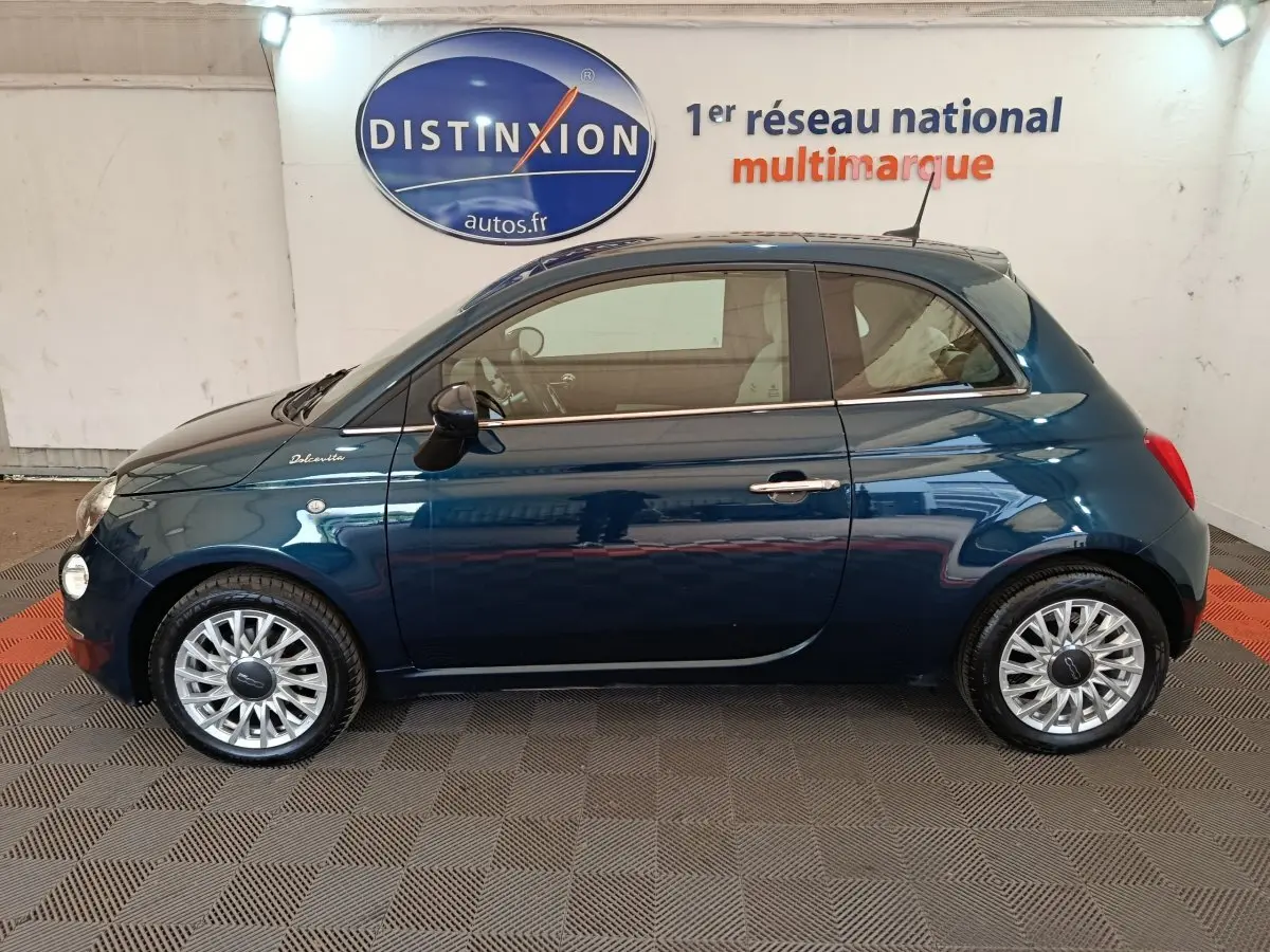 Profil côté gauche d'une Fiat 500 Dolcevita hybride bleu foncé avec jantes alliage et toit noir en intérieur showroom.