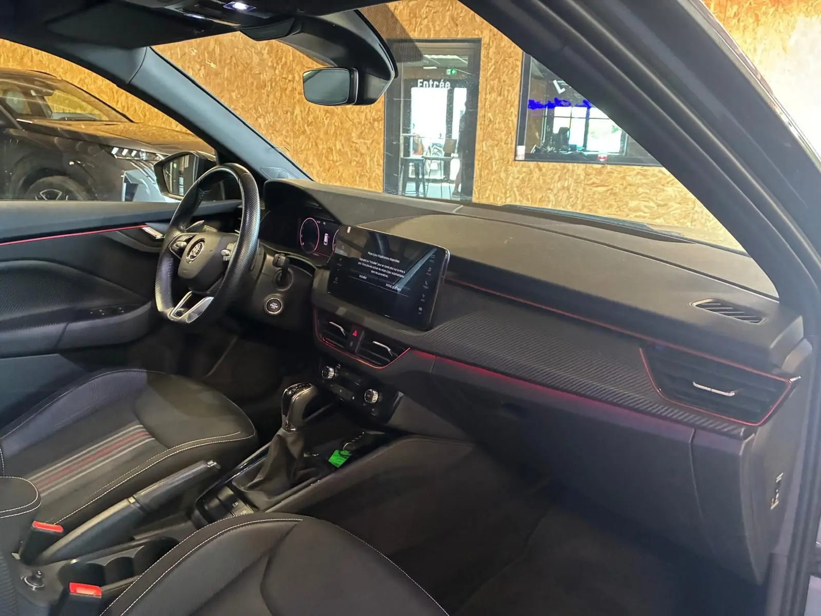 Vue intérieure côté conducteur du Skoda Kamiq 2024 gris foncé, avec tableau de bord noir et éclairage d'ambiance rouge.