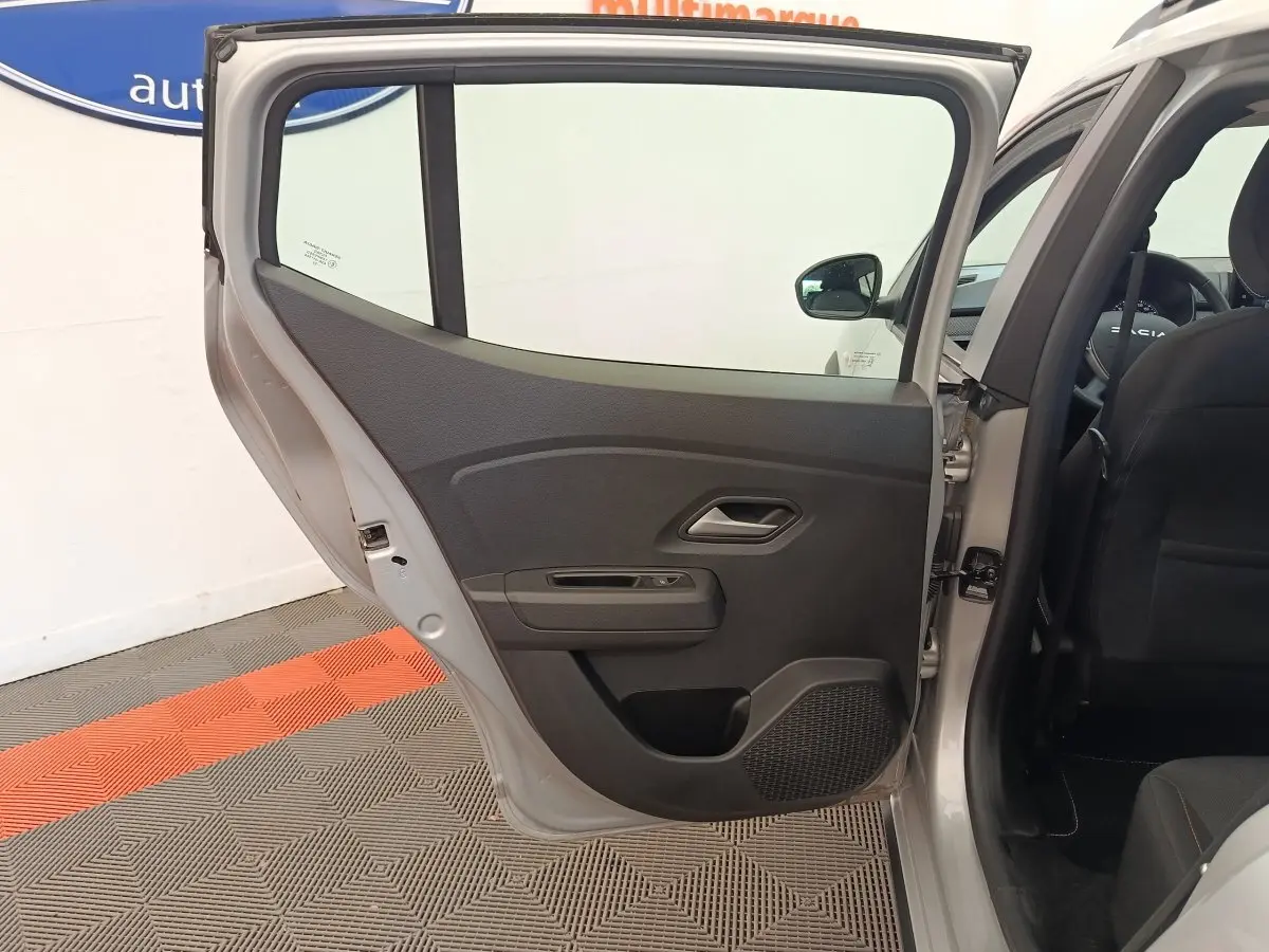 Vue côté gauche sur la porte arrière ouverte d'une Dacia Sandero gris clair, montrant l'intérieur noir et la poignée de porte.