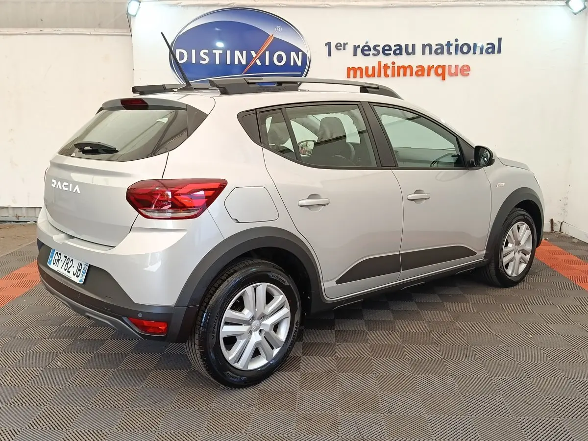 Vue 3/4 arrière droite d'une Dacia Sandero gris clair 2023 avec barres de toit et protections noires sur les bas de caisse.