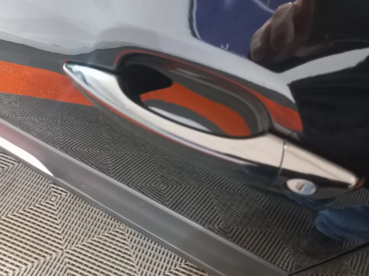 Gros plan sur la poignée de porte chromée d'une Citroën C3 noire, reflet orange et sol texturé visible.