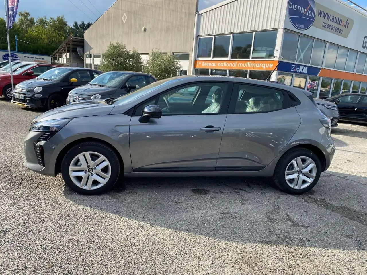 Profil côté gauche d'une Renault Clio 2025 gris schiste garée devant un bâtiment commercial.