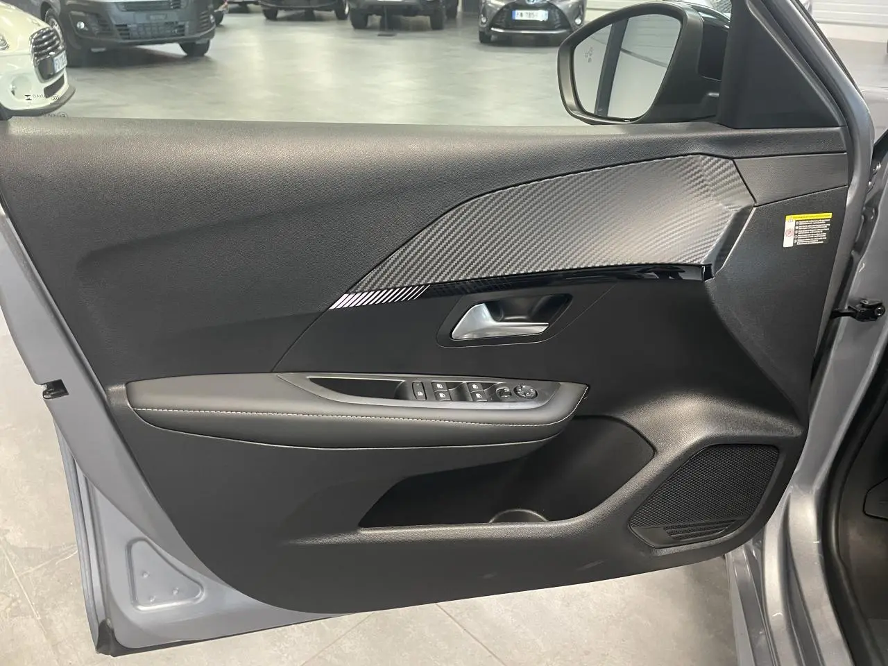 Intérieur côté gauche de la porte avant gris Artense de la Peugeot 208 avec insert carbone et commandes électriques.