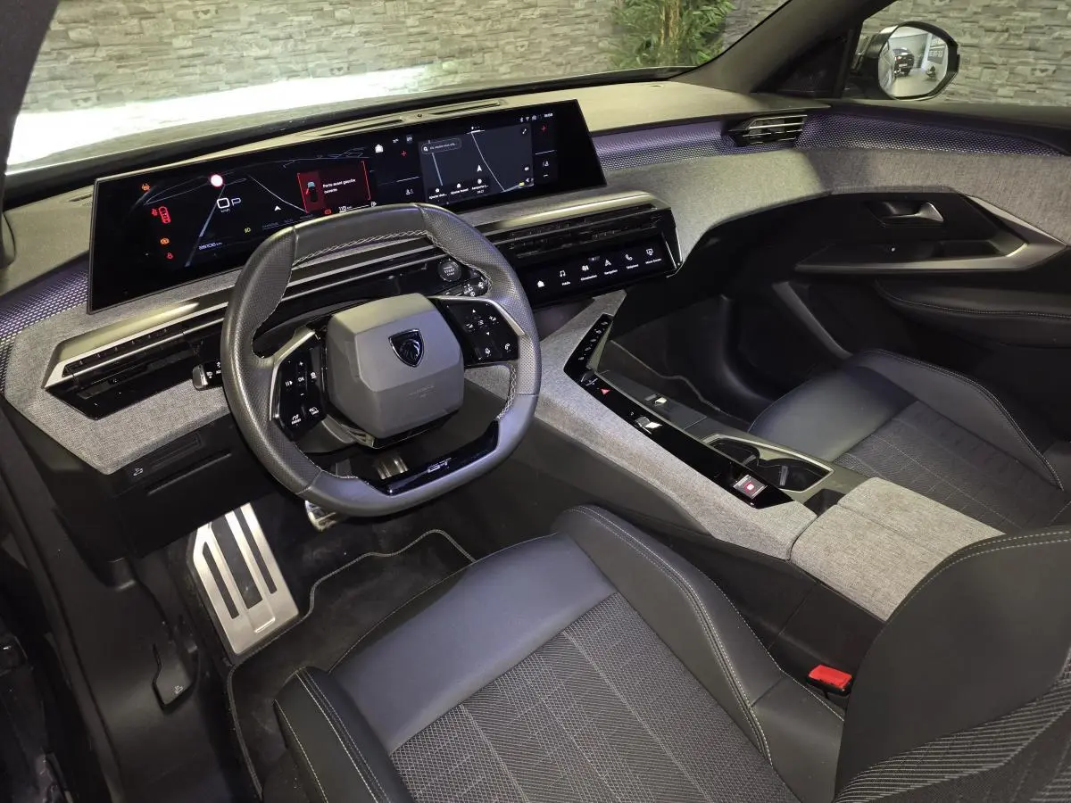 Vue intérieure avant droite du Peugeot 5008 2025, volant compact noir, tableau de bord numérique panoramique et sellerie tissu gris.