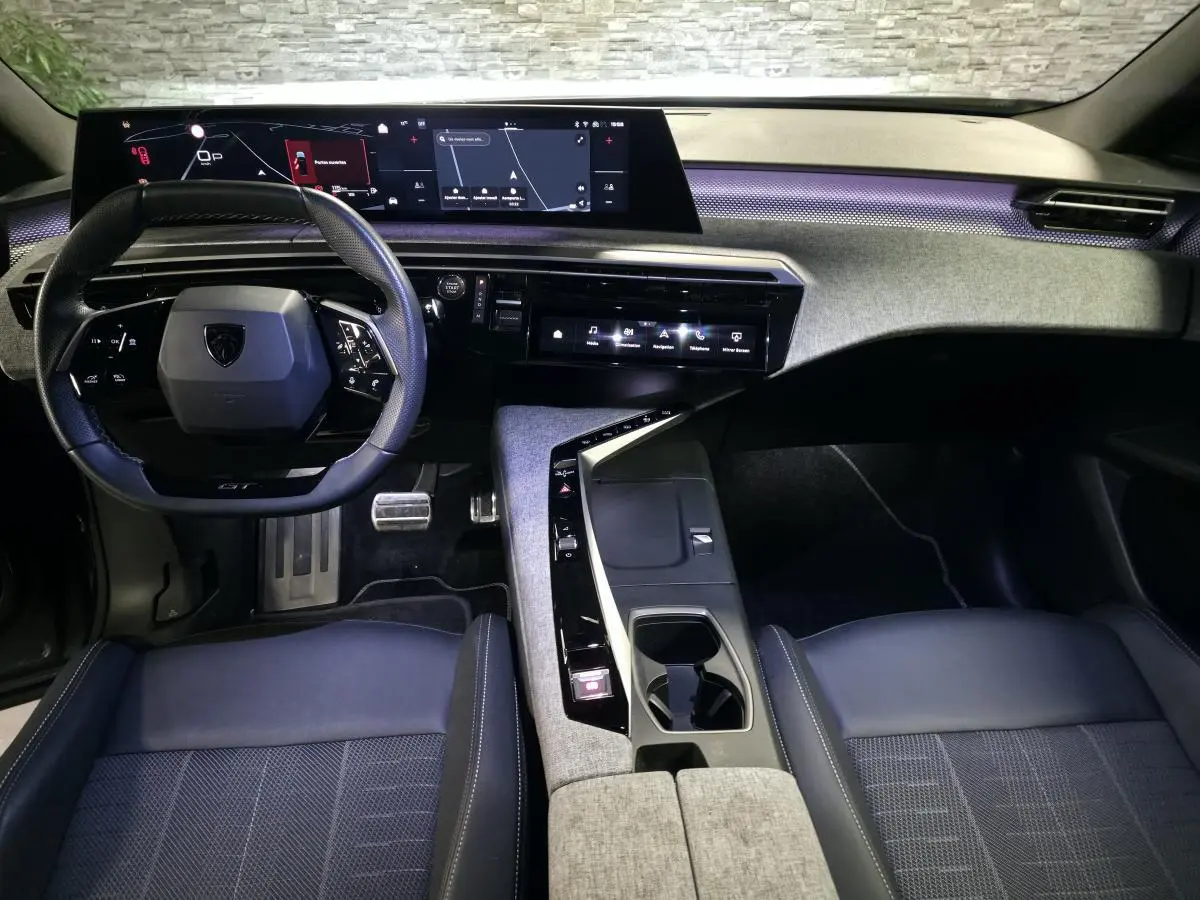 Vue intérieure avant du Peugeot 5008 2025, tableau de bord avec écran panoramique et volant compact noir, ambiance tissu gris et éclairage violet.