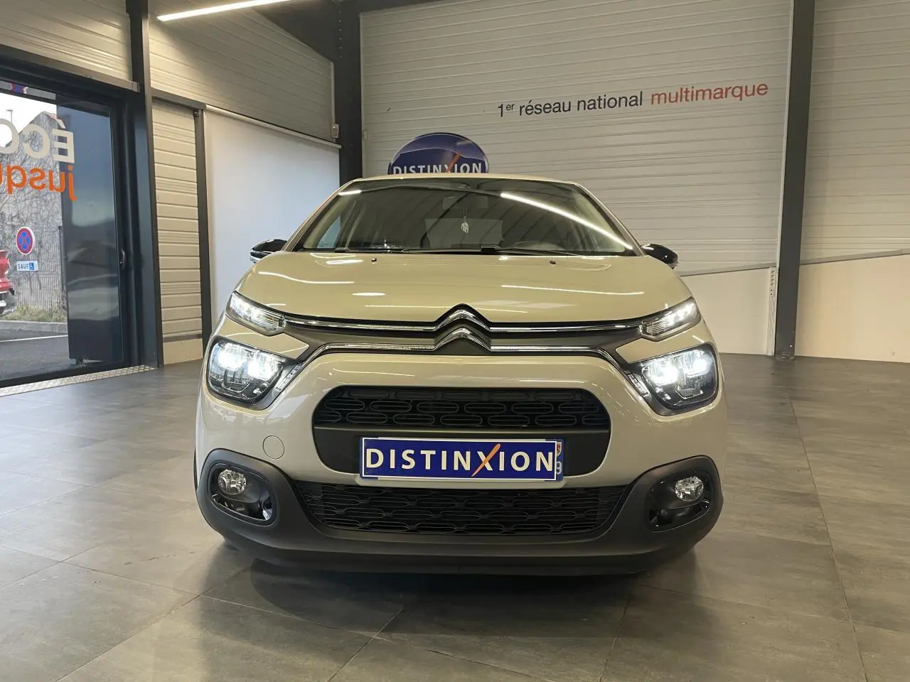 Vue frontale d'une Citroën C3 beige clair 2024 avec feux allumés et calandre noire dans un showroom.
