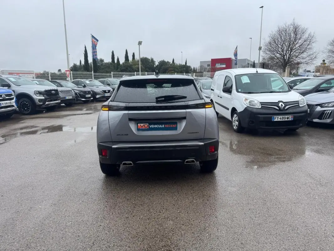 Vue arrière d’un Peugeot 2008 gris avec feux LED allumés, garé sur un parking humide entouré d’autres véhicules.